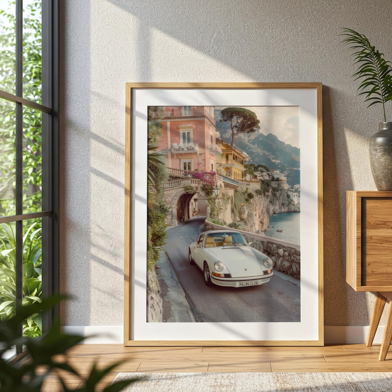 May include: A framed wall art print featuring a white vintage sports car driving on a coastal road. The scene includes pastel-colored buildings, a stone archway, and a cliffside overlooking the sea. The car has a license plate reading PALMO CLUB.