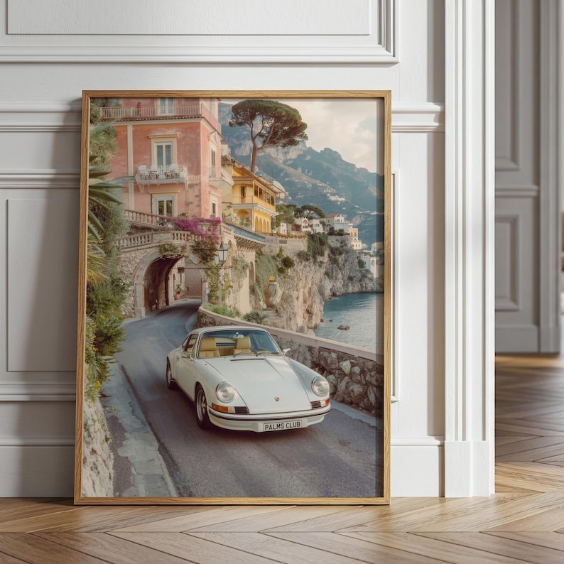 May include: A framed wall art print depicting a white vintage sports car driving on a coastal road. The scene features a pink building with an arched stone tunnel, lush greenery, and a mountain landscape overlooking the sea. The car has a license plate reading PALMS CLUB.