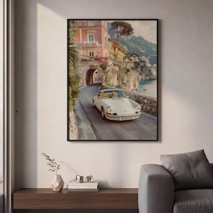 May include: A framed wall art print depicting a white vintage sports car driving on a coastal road. The scene features a stone archway, pastel-colored buildings, and cliffs overlooking the sea. The car license plate reads PALMS CLUB.
