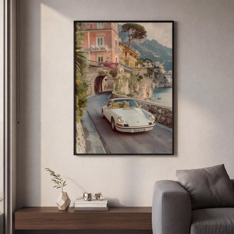 May include: A framed wall art print depicting a white vintage sports car driving on a coastal road. The scene features a stone archway, pastel-colored buildings, and cliffs overlooking the sea. The car license plate reads PALMS CLUB.
