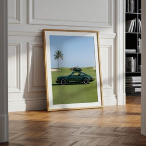 Puede incluir: Impresi&oacute;n enmarcada que muestra un coche deportivo Porsche verde oscuro en un campo de golf. El coche tiene una baca en el techo. Una palmera y cielo azul en el fondo. El marco es de madera clara.