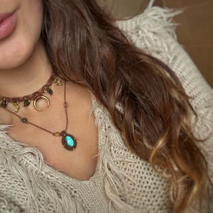 Puede incluir: Primer plano de dos collares hechos a mano. El collar superior es marrón con detalles dorados y un colgante de media luna. El collar inferior tiene un colgante de piedra ovalada azul-verdosa. Los collares se llevan sobre un suéter crema con flecos.