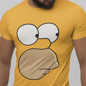 Könnte beinhalten: Senfgelbes T-Shirt mit einem Cartoon-Gesicht. Das Gesicht hat große weiße Augen und einen hellbraunen Mund. Das T-Shirt hat kurze Ärmel und einen Rundhalsausschnitt, mit einem vereinfachten Charakter.