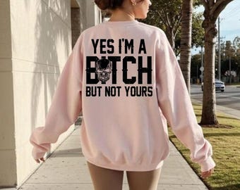 May include: A light pink sweatshirt with the text "YES I'M A BITCH BUT NOT YOURS" in black. The word "BITCH" features a skull graphic. The sweatshirt is a casual style.