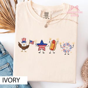 May include: An ivory-colored t-shirt featuring a graphic print of a bald eagle wearing a top hat, a star, a hot dog, and a firework, all with arms and legs. The eagle holds an American flag, while the other characters hold sparklers.