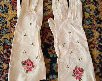 May include: A pair of off-white leather gloves with floral embroidery. Each glove features a large embroidered pink rose with green leaves and smaller floral accents. The gloves are laid out on a patterned rug.
