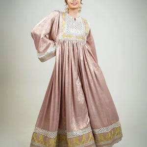 May include: A blush pink dress with long sleeves and a flowing skirt. The dress features intricate gold and white embroidery around the neckline, cuffs, and hem. The fabric has a subtle sheen, and the dress is gathered at the waist.