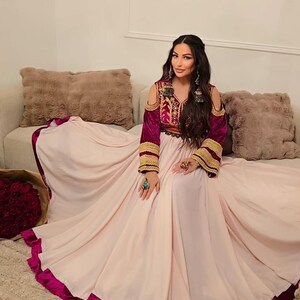 Puede incluir: Un vestido rosa claro con un ribete magenta oscuro. El vestido presenta un corpiño con intrincados bordados en tonos rojos, dorados y morados. Las mangas son largas y moradas con ribete dorado. El vestido es de estilo tradicional.