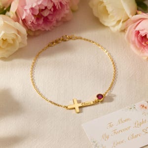 May include: A gold-colored bracelet with a cross design and a small red gemstone. The bracelet has the name "Clare" engraved on it. A card with floral accents reads "To Mom, My Forever Light, Love, Clare".