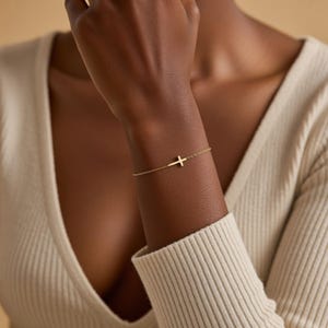 Peut inclure: Un bracelet doré avec une fine chaîne et un pendentif en forme de croix latérale. La croix est positionnée horizontalement sur le poignet. Le bijou est simple et élégant, adapté au quotidien. Le bracelet est en or.