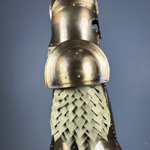 May include: A bronze-colored suit of armor, featuring a helmet, shoulder guards, and a scale-like skirt. The armor has a polished finish and is designed with intricate details, suggesting a historical or fantasy theme.
