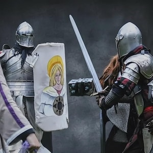 May include: Two knights in full armour face each other, one holding a long sword and the other a shield. The shield features a painted image of a woman with a yellow halo. The knights are in a historical reenactment.