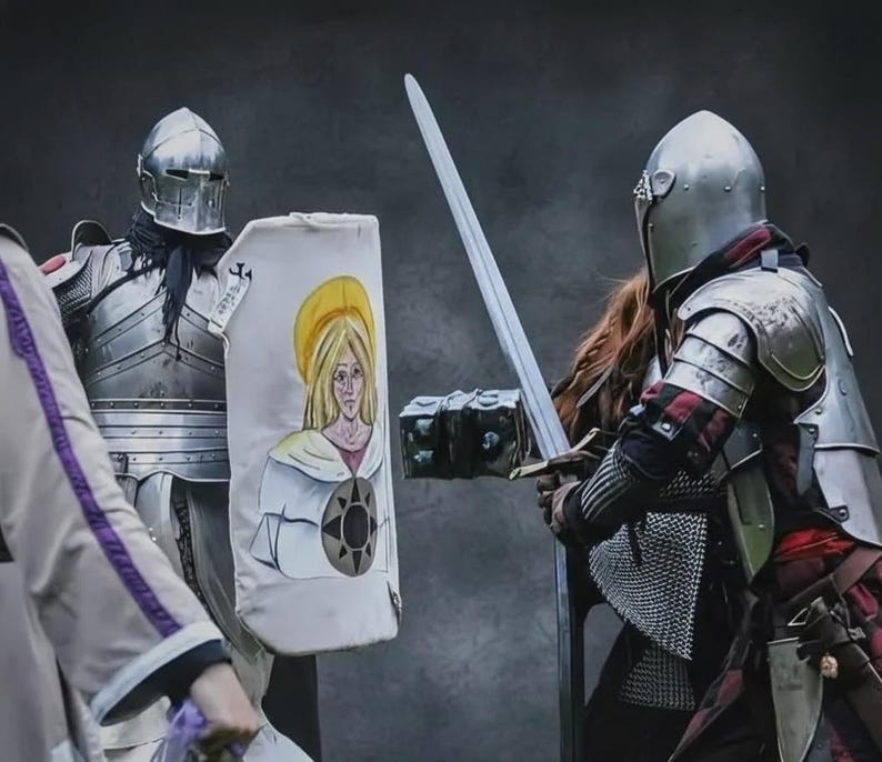 May include: Two knights in full armour face each other, one holding a long sword and the other a shield. The shield features a painted image of a woman with a yellow halo. The knights are in a historical reenactment.