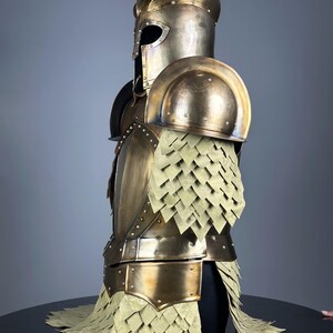 May include: A full suit of antique gold-colored armor, including a helmet, shoulder pads, and chest piece. The armor features riveted construction and a scale-like skirt. The helmet has a visor, and the armor is displayed on a black surface.