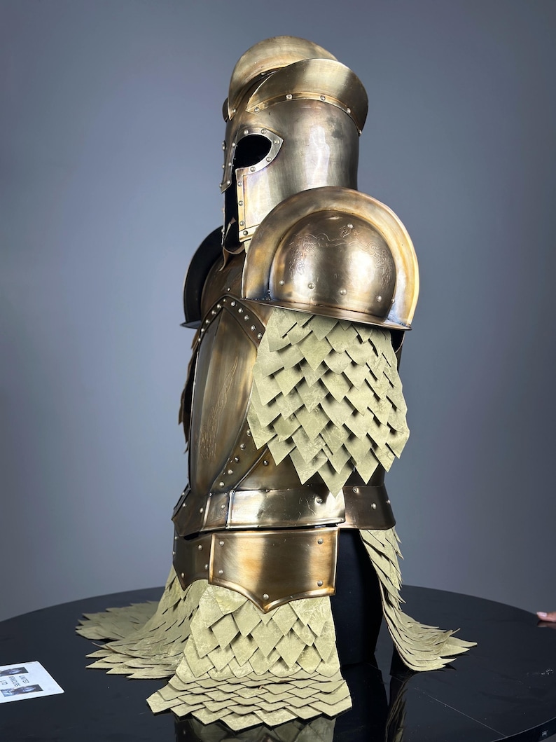 May include: A full suit of antique gold-colored armor, including a helmet, shoulder pads, and chest piece. The armor features riveted construction and a scale-like skirt. The helmet has a visor, and the armor is displayed on a black surface.