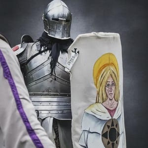 May include: A suit of medieval armour with a polished steel helmet and a shield. The shield features a painted image of a woman with a golden halo and a brown emblem. The armour is worn by a person.