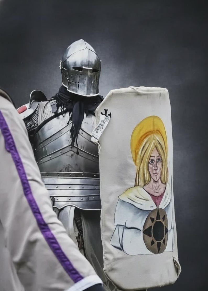 May include: A suit of medieval armour with a polished steel helmet and a shield. The shield features a painted image of a woman with a golden halo and a brown emblem. The armour is worn by a person.
