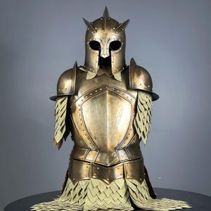 May include: A bronze-colored suit of armor, featuring a helmet with pointed accents and a visor. The chest plate and shoulder guards are detailed with rivets. The armor includes layered, scale-like elements around the shoulders and waist, creating a medieval aesthetic.