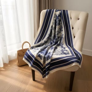 Puede incluir: Un pañuelo cuadrado de seda drapeado sobre una silla. El pañuelo presenta un fondo crema con un diseño de paisaje azul y blanco. Tiene un borde azul marino con rayas. Un bolso de mano marrón claro está cerca.