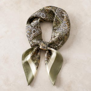 May include: Olive green paisley patterned scarf with cream-colored stripes on the ends. The scarf is knotted in the center, with the ends hanging down. The scarf has a silky sheen and a detailed floral design.