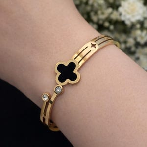 Könnte beinhalten: Goldfarbenes Armband mit einem schwarzen, kleeblattförmigen Mittelstück und zwei klaren, runden Akzentsteinen. Das Armband hat ein Doppelband-Design mit Stern- und Liniendetails. Ein stilvolles Schmuckstück.