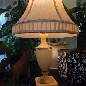 May include: A cream-colored table lamp with a beige lampshade. The lampshade has a scalloped edge and a brown trim. The base of the lamp is a light beige color with a square base. The lamp is on a black surface.