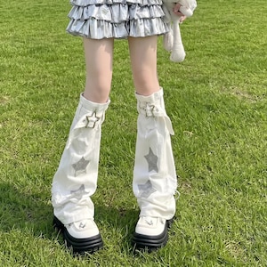 May include: White leg warmers with star embellishments and silver buckles. The leg warmers are paired with white platform shoes with black soles. The background is green grass.