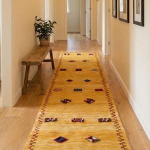 Puede incluir: Una alfombra larga y rectangular de color amarillo con un patrón geométrico en un pasillo. La alfombra tiene un borde rojo en zigzag y presenta varias formas en rojo, azul y blanco. El pasillo tiene paredes de color claro y suelo de madera.