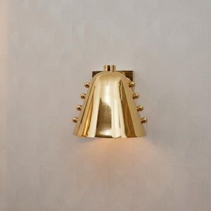 Op de afbeelding: Een goudkleurige wandlamp met een klokvormige kap en een vierkante montageplaat. De kap is versierd met kleine, ronde decoratieve elementen. De lamp is ontworpen om aan de muur te worden bevestigd en zorgt voor sfeerverlichting.