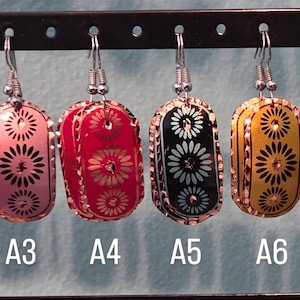 May include: A collection of eight colorful earrings with a rectangular shape. Each earring features a decorative floral design and a silver-colored hook. The earrings are in various colors, including copper, green, pink, red, black, yellow, purple, and blue.