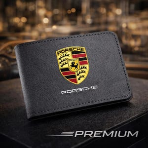 May include: A black bi-fold wallet featuring the Porsche logo in gold, red, and black on the front. The word PORSCHE is printed in white below the emblem. The wallet has visible black stitching along the edges and a textured surface finish.