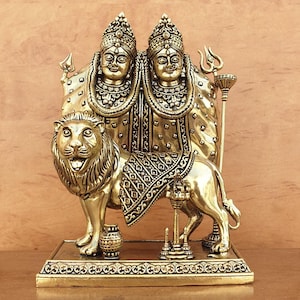 Pode incluir: Estátua de latão dourado retratando duas figuras com toucados ornamentados e roupas detalhadas. As figuras são ladeadas por um leão, com padrões intrincados em sua sela. A estátua é montada em uma base decorativa, mostrando um tema religioso.