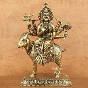 Peut inclure: Une statue en métal doré représentant une divinité aux bras multiples assise sur une chèvre. La figure porte une couronne et des bijoux ornés, tenant un trident et une masse. La base rectangulaire présente des motifs floraux et le texte Kalyan Arts.