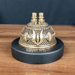 May include: A gold-toned metal dome-shaped object featuring a pair of feet on top, intricate cross-hatched patterns, and a central trident-like symbol. The object is mounted on a circular black base with the text Kalyan Arts printed on the front.