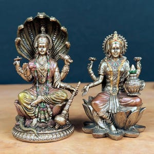 Op de afbeelding: Twee gedetailleerde bronstinten beelden van goden. Eén heeft een veelkoppige slangenhoofdtooi, de andere zit op een lotusbloem. Beide figuren zijn versierd met ingewikkelde sieraden en houden symbolische voorwerpen vast. De beelden staan op een houten oppervlak.