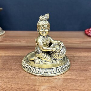 Può includere: Una statuetta in ottone raffigurante un bambino inginocchiato su una base circolare a forma di loto, che tiene in mano un piccolo vaso. La figura presenta gioielli dettagliati, una piuma di pavone tra i capelli e la scritta Kalyan Arts.