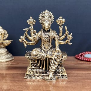 May include: A brass-colored metal statue of the deity Varaha, depicted with a boar head and multiple arms, seated on an ornate rectangular base. The figure wears detailed jewelry and garments, with hands holding various symbolic objects.