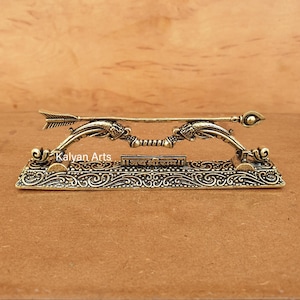 May include: A metallic gold-toned decorative bow and arrow set resting on a rectangular base with intricate scrollwork patterns. The centre of the base features a small plaque with the inscription "जय श्री राम" and the text "Kalyan Arts" is visible.