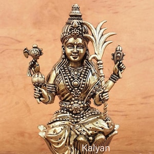 Può includere: Una statuetta in ottone color oro raffigurante una divinità a quattro braccia seduta su una base di loto. La figura indossa una corona ornata, collane a strati e gioielli, tenendo un gambo di canna da zucchero e una mazza. La parola Kalyan è visibile.