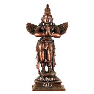 Puede incluir: Una estatua de metal de color bronce de una figura alada de pie sobre una base rectangular. La figura tiene las manos juntas en posición de oración, viste ropa tradicional y presenta el texto Kalyan Arts en la parte frontal de la base.
