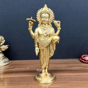 May include: A polished brass statue of a four-armed deity standing on a circular base. The figure wears traditional draped garments, jewelry, and a halo-like crown, holding a small lamp and a vessel. The text Kalyan Arts is visible at the base.