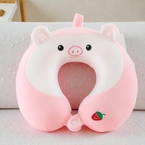 Pode incluir: Uma almofada de viagem rosa em forma de porco, com rosto e orelhas brancas. A almofada em forma de U tem um bordado de morango. O porco tem olhos pretos e nariz rosa. Projetada para suporte do pesco&ccedil;o, mede cerca de 30 cm de largura.