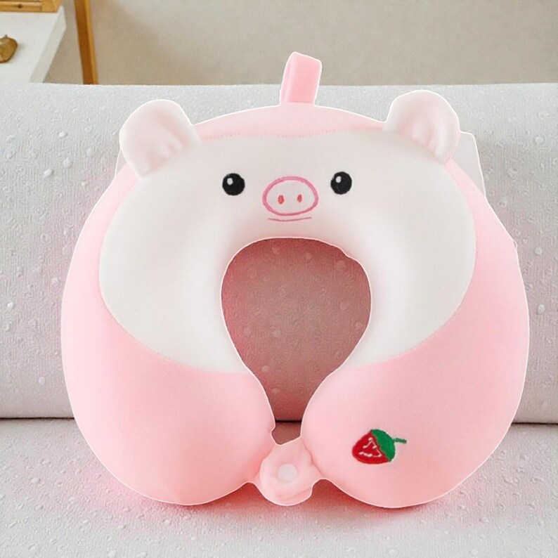 Pode incluir: Uma almofada de viagem rosa em forma de porco, com rosto e orelhas brancas. A almofada em forma de U tem um bordado de morango. O porco tem olhos pretos e nariz rosa. Projetada para suporte do pesco&ccedil;o, mede cerca de 30 cm de largura.