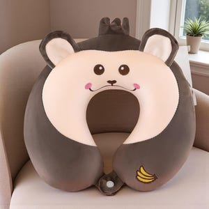 Pode incluir: Uma almofada de viagem de pel&uacute;cia em forma de rosto de macaco. O rosto &eacute; bege claro com olhos castanhos e bochechas rosadas. Uma pequena banana bordada est&aacute; na lateral. A almofada foi projetada para apoiar o pesco&ccedil;o.