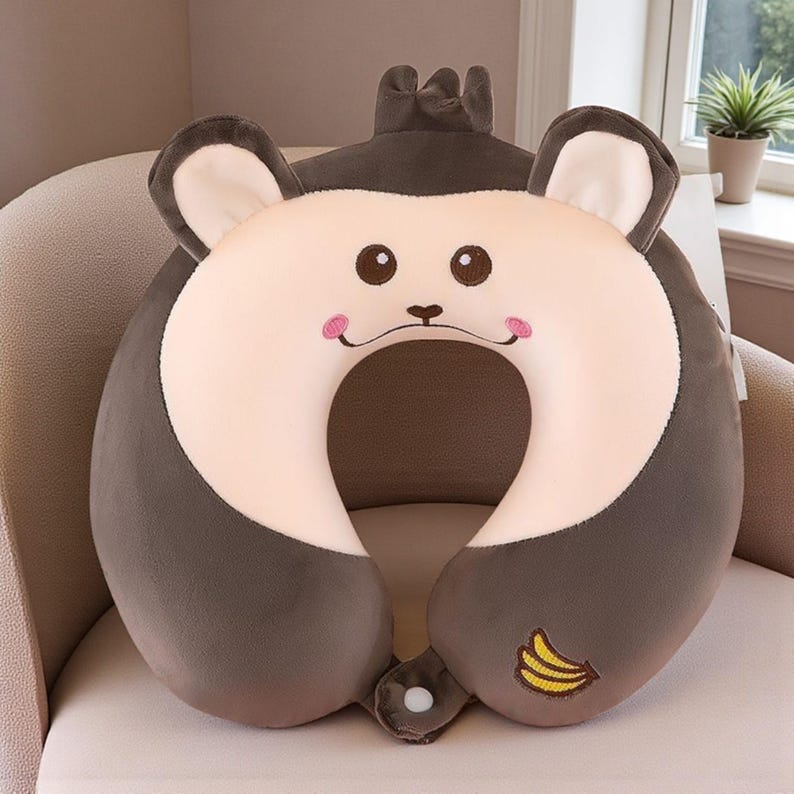 Pode incluir: Uma almofada de viagem de pel&uacute;cia em forma de rosto de macaco. O rosto &eacute; bege claro com olhos castanhos e bochechas rosadas. Uma pequena banana bordada est&aacute; na lateral. A almofada foi projetada para apoiar o pesco&ccedil;o.