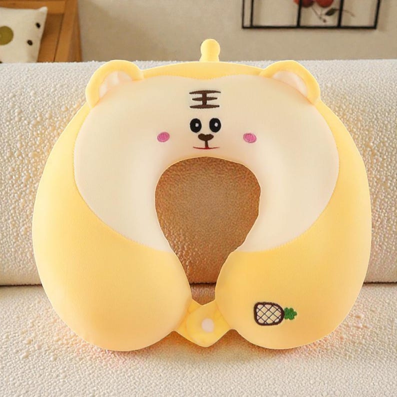 Pode incluir: Almofada de viagem de pel&uacute;cia amarela em forma de cabe&ccedil;a de tigre. A almofada em forma de U tem uma abertura central. O rosto do tigre apresenta olhos pretos, um nariz e o caractere chin&ecirc;s para "rei". Um pequeno abacaxi &eacute; bordado na lateral.