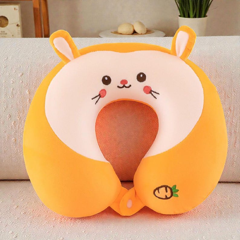 Pode incluir: Uma almofada de viagem de pel&uacute;cia laranja em forma de coelho. A almofada tem um centro branco com um rosto de coelho de desenho animado, incluindo olhos pretos, bigodes e um pequeno nariz vermelho. Possui duas orelhas verticais e um &iacute;cone de cenoura.