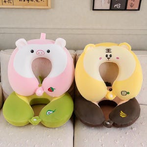 Pode incluir: Duas almofadas de viagem de pel&uacute;cia. Uma &eacute; rosa e branca, em forma de porco com base verde. A outra &eacute; amarela e marrom, em forma de tigre. Ambas t&ecirc;m um design em forma de U para suporte do pesco&ccedil;o e s&atilde;o projetadas para viagens.