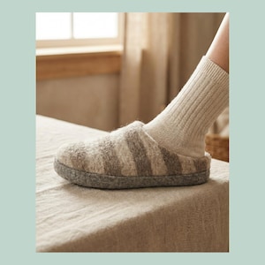 May include: A close-up of a grey and beige striped slipper with a grey sole. A person is wearing a beige sock inside the slipper. The slipper is made of a soft, textured material.