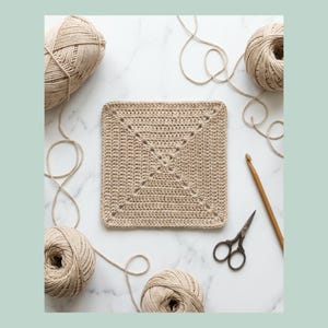 May include: A square, beige crochet piece with a geometric design is surrounded by beige yarn balls and loose yarn strands. A wooden crochet hook and small scissors are also present, suggesting a crafting theme.